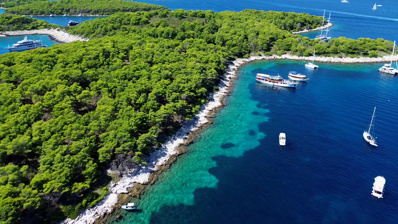 Un drone scende su una costa bianca con una lussureggiante foresta verde di un'isola croata, con barche ancorate nelle vicinanze nel mare calmo e turchese.