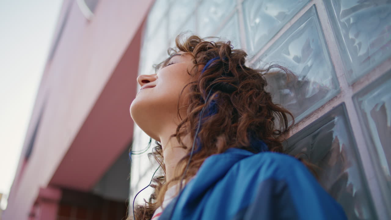 mujer positiva disfrutando de la música en auriculares apoyándose en la pared de vidrio al aire libre en primer plano