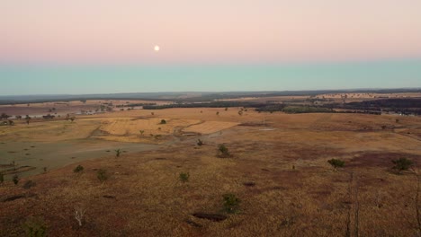 Early-daytime-Full-moon-over-stretched-brownish-yellow-grassy-plains,-aerial-view