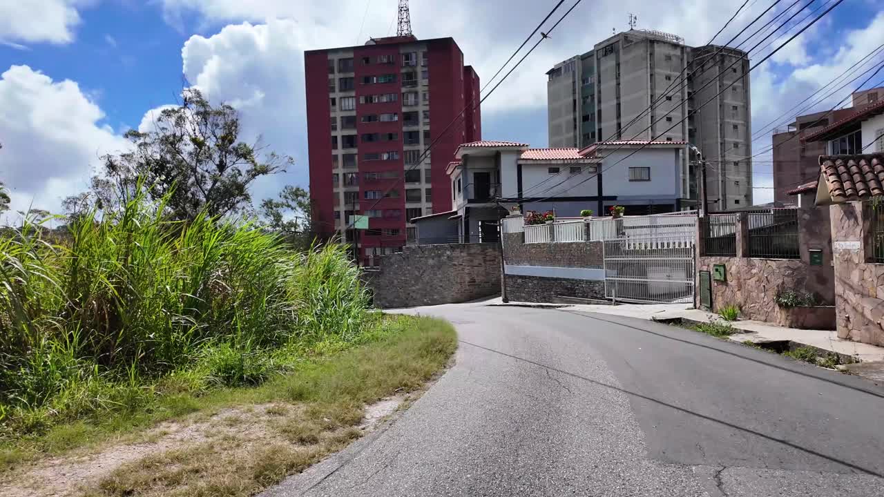 Plano aberto de uma estrada sinuosa e tranquila em San Antonio, subúrbio de Caracas, mostrando uma mistura de casas tradicionais com telhados vermelhos e edifícios modernos de tijolos, cercados por vegetação.