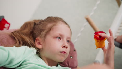 Cute girl relaxing indoors eating juicy red apple almost finished looking thoughtfully at remaining fruit with partial view of another child nearby