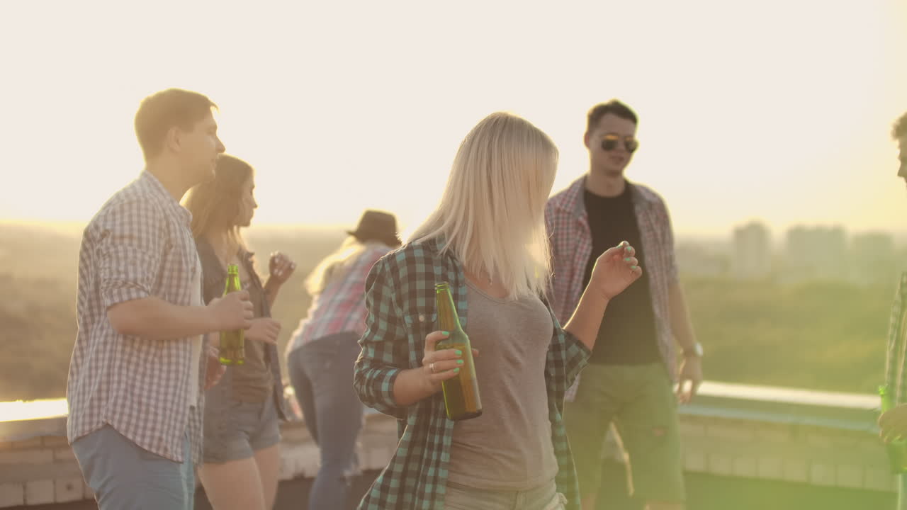 The American blonde woman is dancing on the roof with her friends who drinks beer on the party. She smiles and enjoys the time she is dancing with a beer in a gray tank top and a dark green plaid shirt.