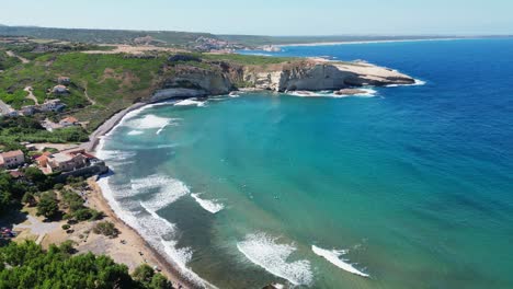 Surfers-in-blue-bay-at-S'-Archittu-beach-town,-west-coast-of-Sardinia---4k-Aerial