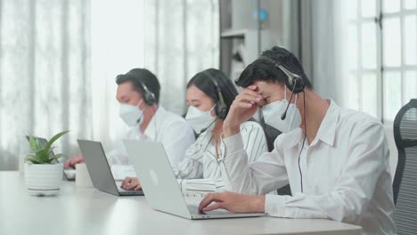 a man of three asian call center agents wearing headset and mask headache to work while two of his colleagues are speaking and typing during the call with customer at the office