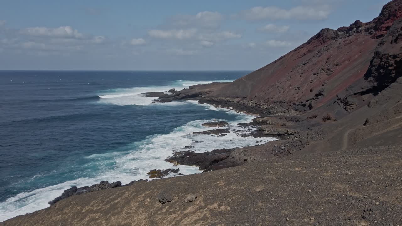 Turquoise ocean waves crash against the dark volcanic rock of the lanzarote coastline, creating white foam under a blue sky dotted with white clouds