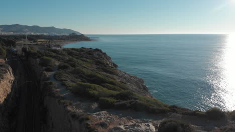 drone pilot flying over himself off cliff as train comes out tunnel underneath and drives towards coastal city passes underneath towards