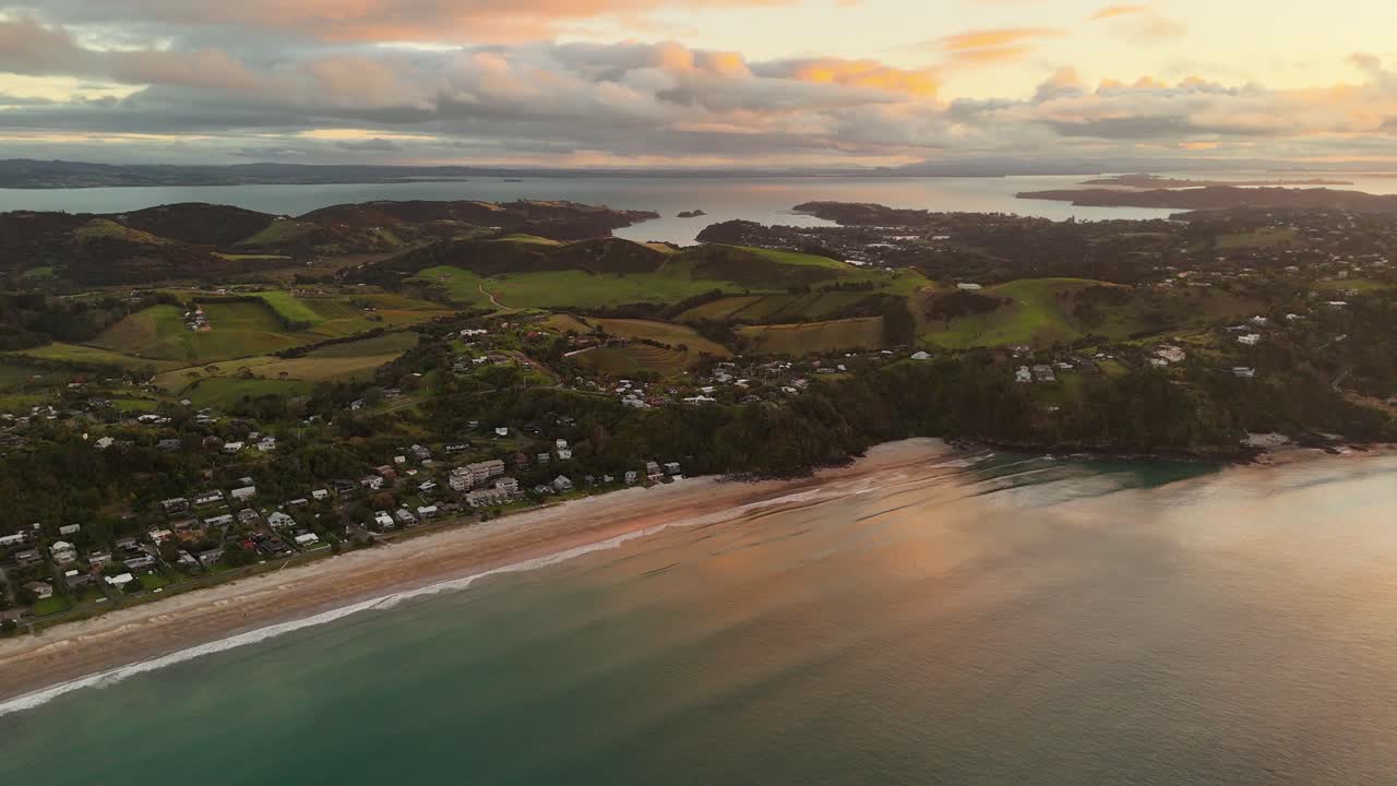Aerial wide shot of Onetangi Beach at golden Sunset. Peaceful scene with idyllic and Tropical hills on Waiheke Island, New Zealand. Colored island with luxury Houses and homes in first row.