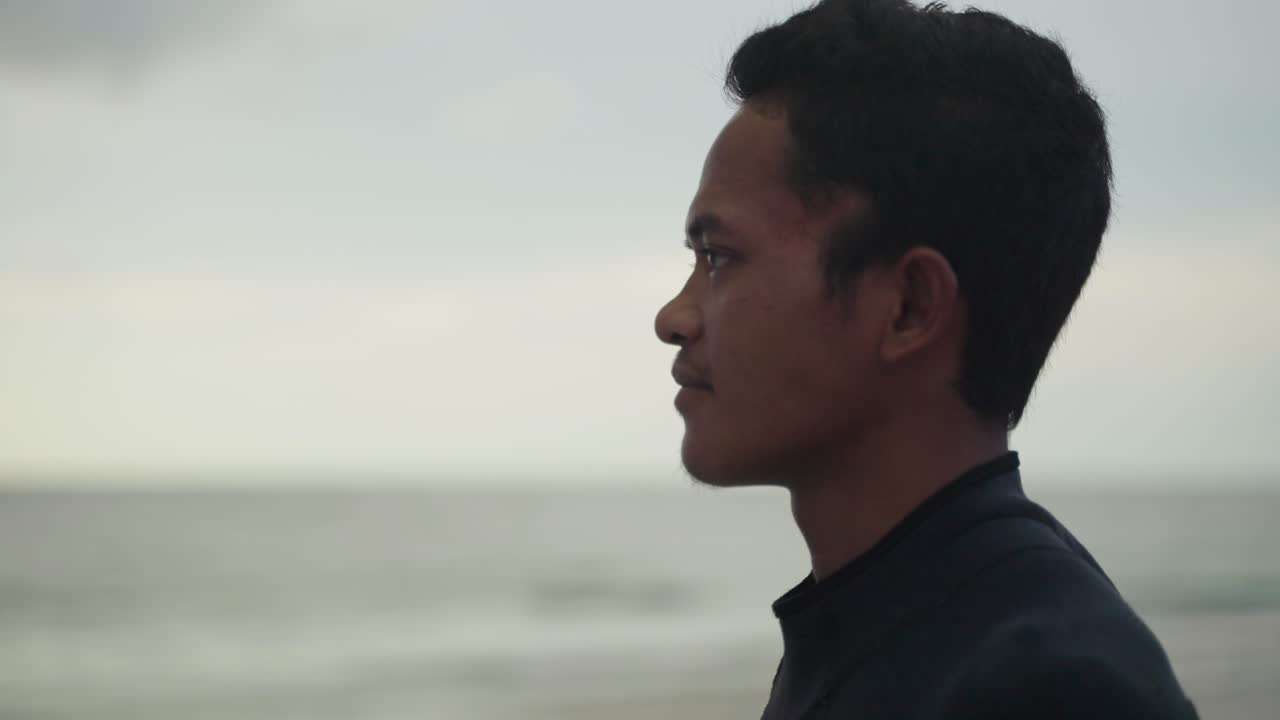 A young male Indonesian man shown in side view profile looking out to the ocean with an out of focus ocean background