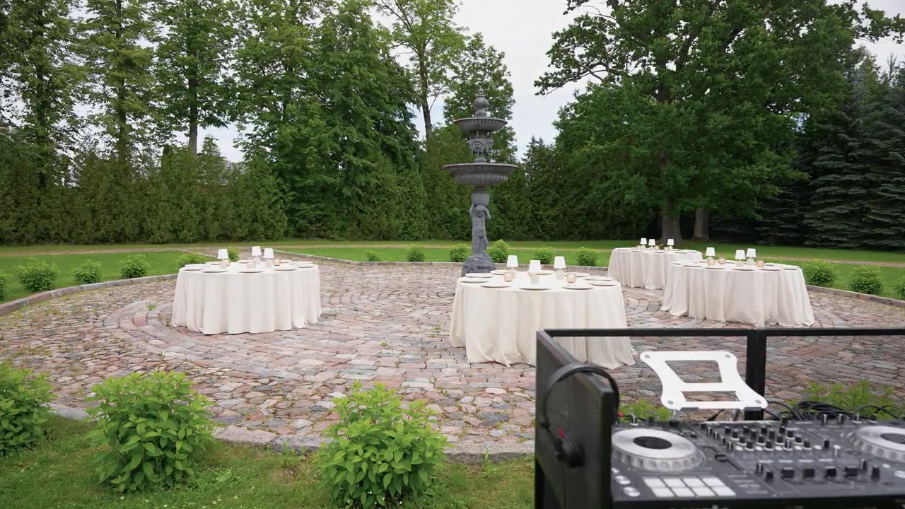 Circular outdoor venue with round tables draped in white linens set around a stone fountain, DJ console in foreground, lush greenery and soft daylight create a refined, festive mood.