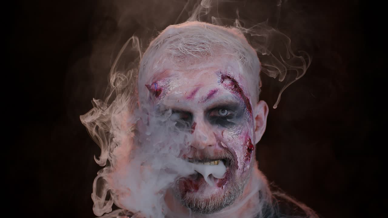 Sinister man horrible scary Halloween zombie looking ominous at camera blows smoke from nose, mouth