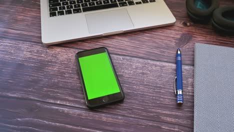 view above on the smartphone with a green screen on the wooden table with a laptop, headphones and office stuff. chroma key