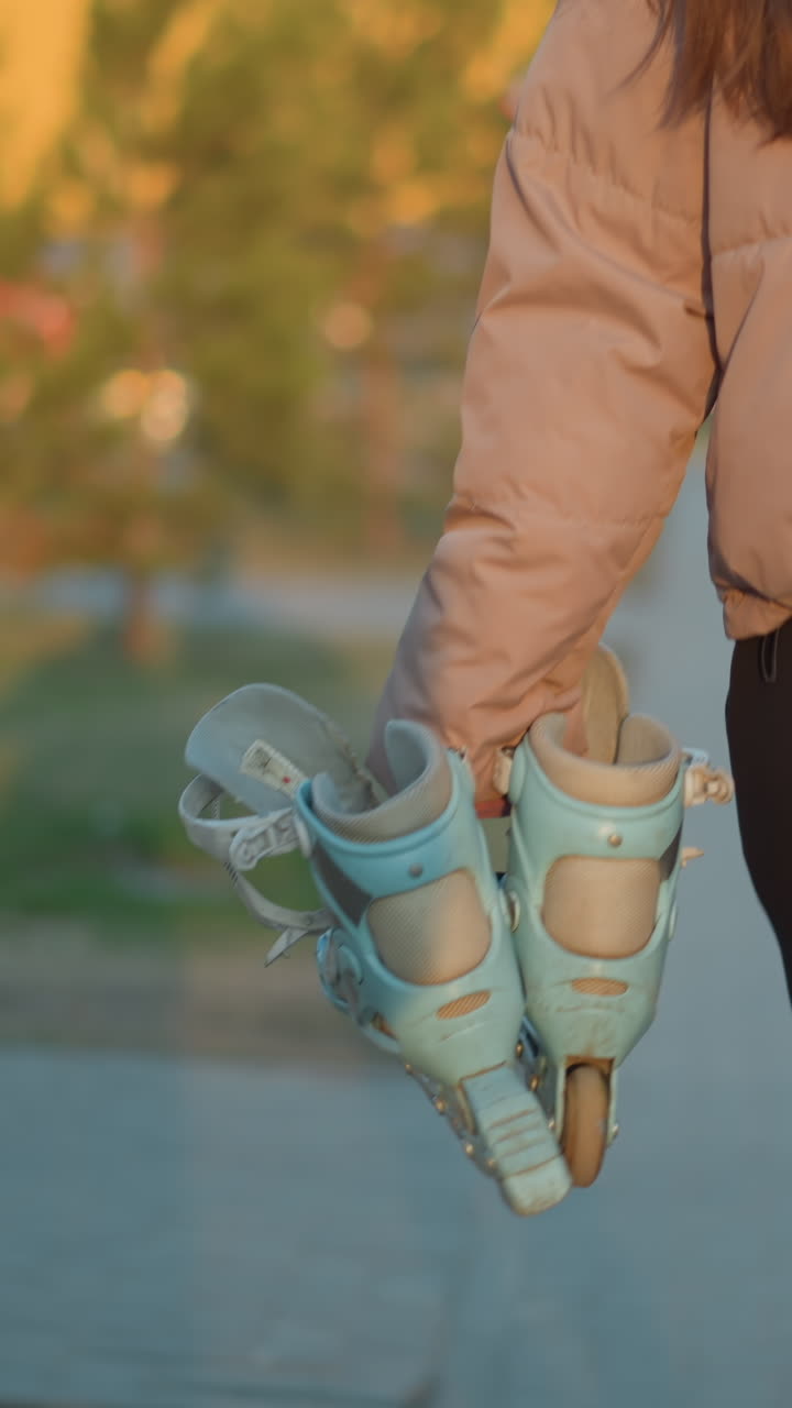 A back shot of a girl dressed in a peach jacket and black trousers, carrying a gym bag and a pair of rollerblades as she walks down a path at sunset