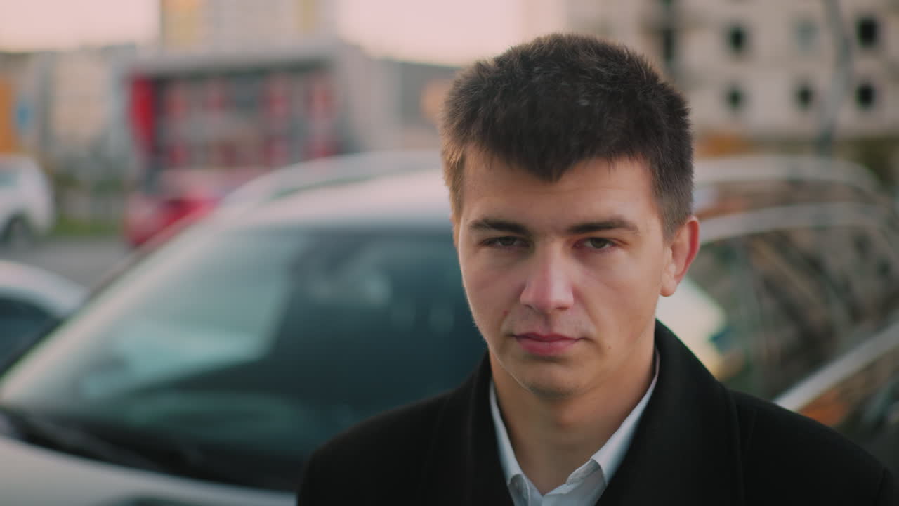Man standing close to car with stern look, blurred background shows parked cars, distant building at dusk, with moving blurred figure in background
