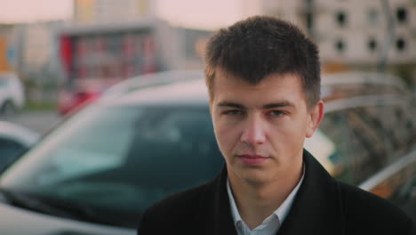 man standing close to car with stern look, blurred background shows parked cars, distant building at dusk, with moving blurred figure in background