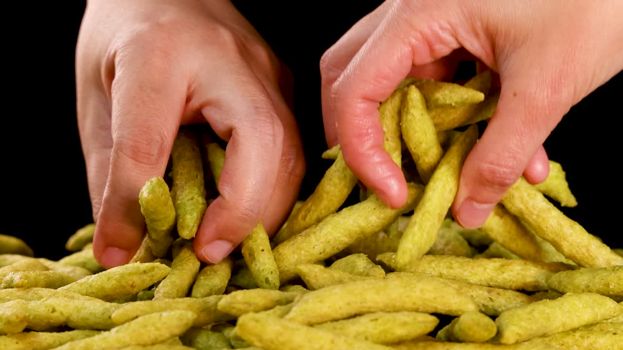 Human hand selects and lifts green pea puffed snacks from pile, studio lighting, black background