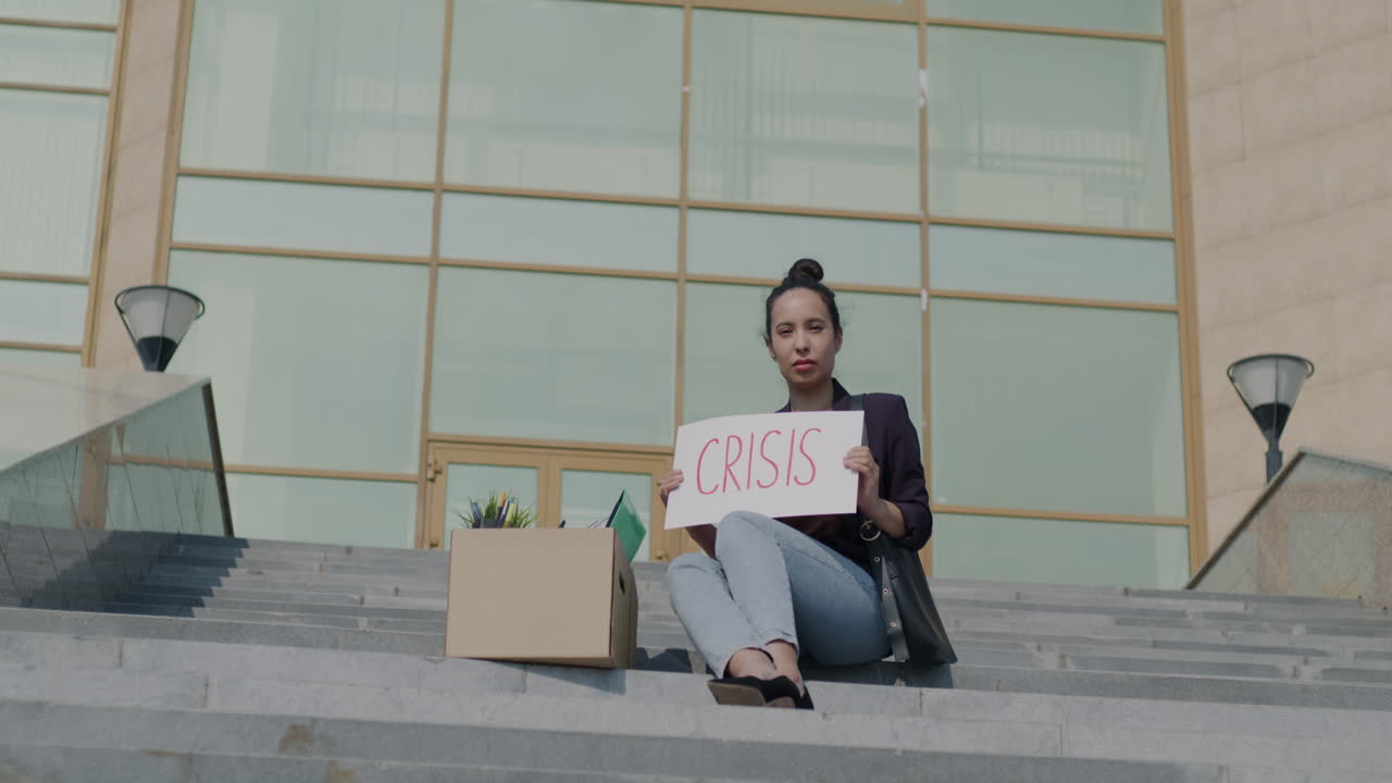 Woman holding a sign saying 'Crisis' sitting on the steps outside an office building