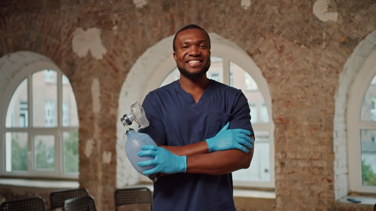 Portrait: A professional Black male doctor in a blue medical uniform stands and smiles against the background of brick walls and large white windows