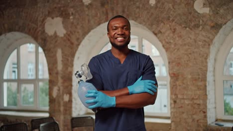 retrato: un médico profesional de raza negra en un uniforme médico azul se para y sonríe contra el fondo de paredes de ladrillo y grandes ventanas blancas
