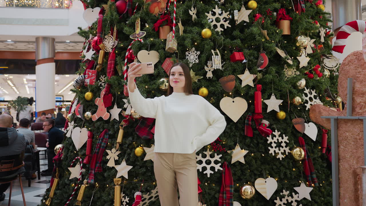 Lady in white top poses with hand on waist taking selfie in front of beautifully decorated Christmas tree inside mall, festive ornaments, golden lights, and seated shoppers create holiday ambiance