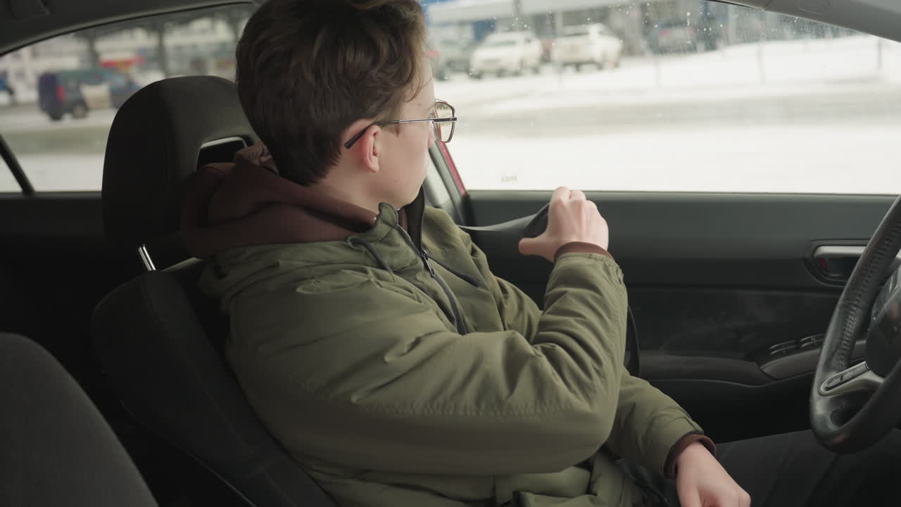 side view young man in green jacket inside car reaching to remove seat belt with focused expression, hand on steering wheel, background through window shows snowy urban street