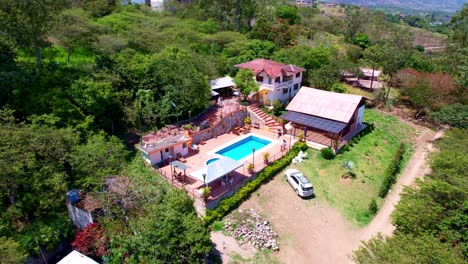 aerial shot with drone from the view of a hacienda with a red roof and white walls surrounded by large leafy and green trees with a blue pool and a sunny summer day