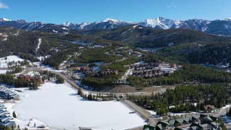 Overhead drone view captures the town of Big Sky, Montana blanketed in snow, with charming vacation condos, frosted roads, and pine-covered hills beneath towering mountain peaks