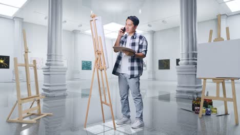 cuerpo lleno de artista masculino asiático hablando en teléfono inteligente mientras pinta en lienzo con pinturas al óleo y pincel en la galería