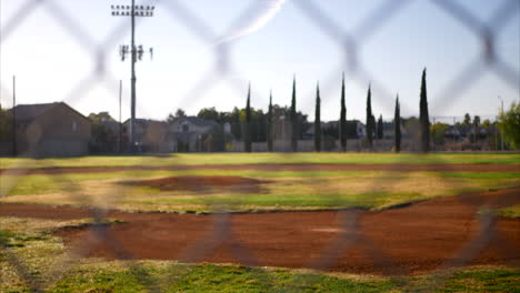 Deslizamiento-Lento-Hacia-La-Izquierda-Mirando-Hacia-Un-Campo-De-Béisbol-Verde-Vacío-Desde-Detrás-De-Una-Cerca-De-Eslabones-De-Cadena-En-Un-Parque-Local-Al-Amanecer