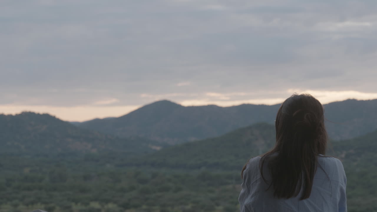 A woman stands still, looking thoughtfully at a mountain range in a peaceful and remote natural setting, conveying solitude, awe, and connection with nature