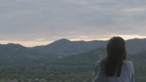 a woman stands still, looking thoughtfully at a mountain range in a peaceful and remote natural setting, conveying solitude, awe, and connection with nature