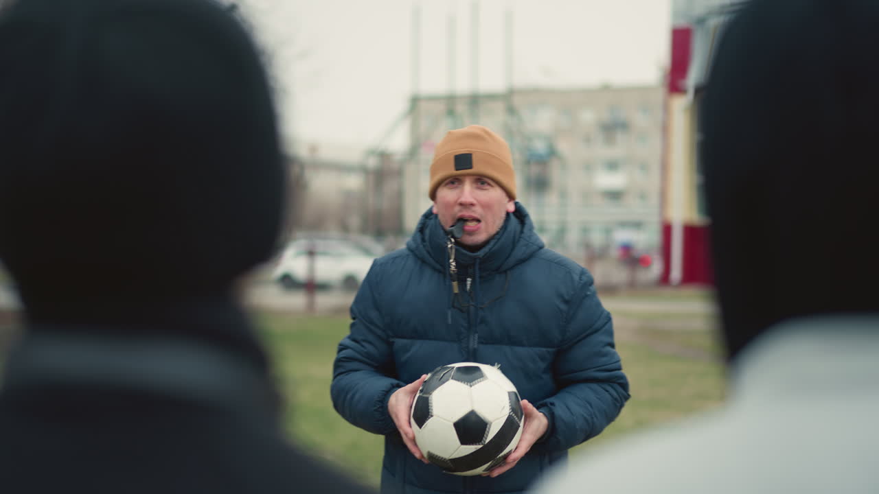 Close-up of a coach instructing two boys with a whistle in his mouth, holding a soccer ball, the coach throws the ball at the person wearing a gray top