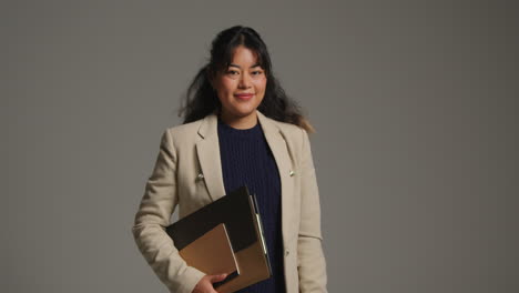 studio portrait of female teacher or businesswoman standing against grey background with folder under her arm