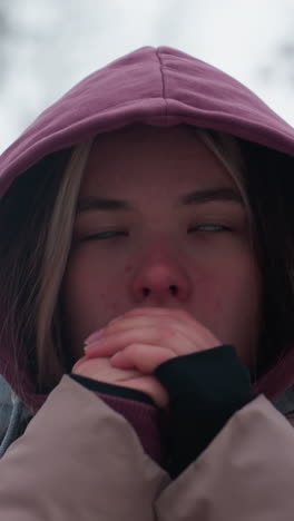 primer plano de una mujer fría ajustando la gorra de la capucha para mantenerse caliente en el frío clima invernal, el aire helado enrojece su cara mientras asegura su capucha, atmósfera invernal borrosa con ramas de árboles desnudas en el fondo