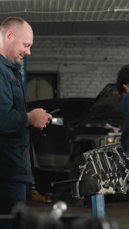 ingenieros mecánicos en un taller, uno trabajando atentamente en un motor con una llave inglesa, de vuelta a la cámara, mientras su colega usa su teléfono, rodeado de equipos mecánicos