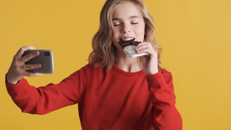 Teenage-Caucasian-girl-eating-chocolate-bar-and-taking-selfies-with-smartphone.