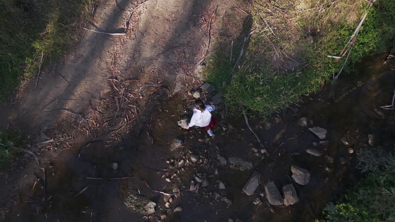 Luftfoto af en pige, der yndefuldt træder over solbeskinnede sten i et vandløb i Warrandyte Park, Melbourne. Det sene aftenlys kaster lange skygger og skaber et fredfyldt naturmaleri.