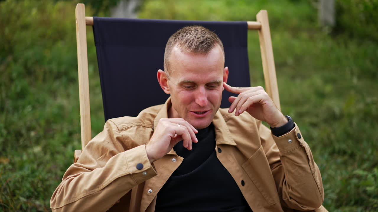 Positive relaxed man looks at the screen of his laptop. Male freelancer sitting comfortably in a garden chair working remotely from nature.