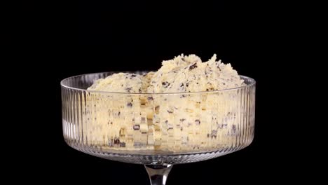 A metal scoop serves vanilla ice cream with chocolate chips and brownie pieces into a clear glass dessert dish against a black background, under studio lighting