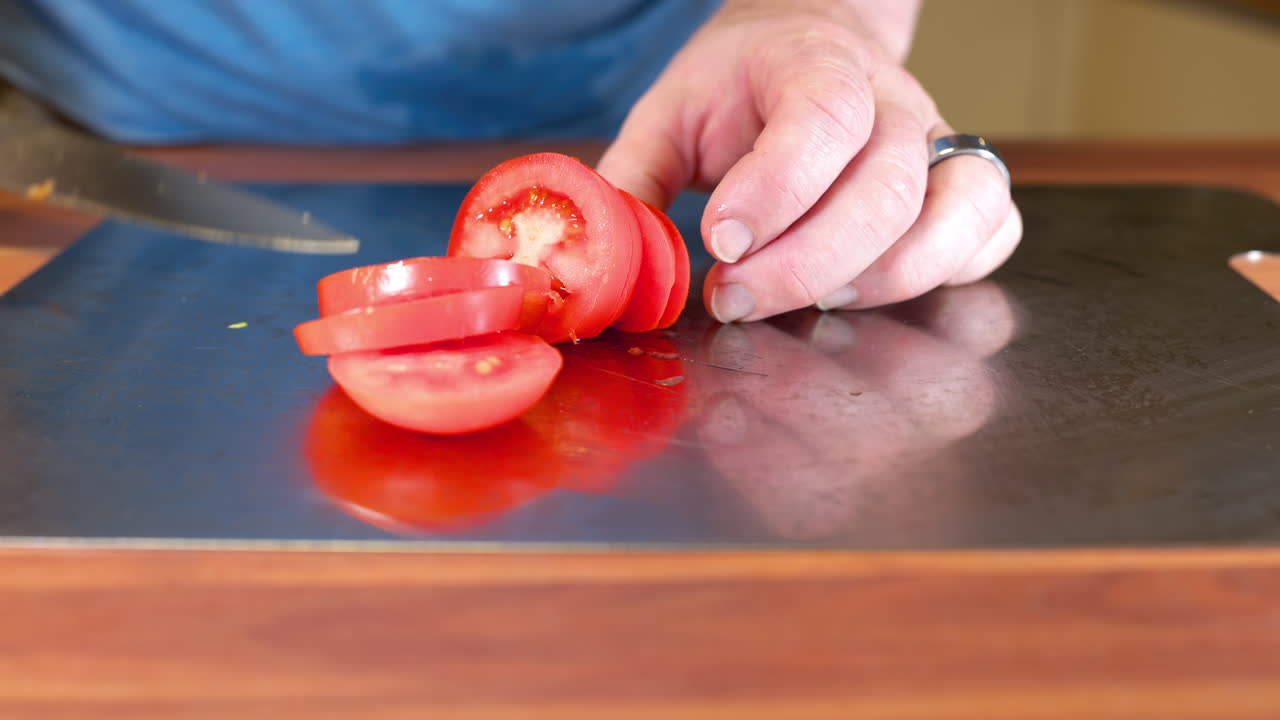 Close-up of a person slicing a fresh tomato on a stainless-steel cutting board in a kitchen. The hands carefully hold a sharp kitchen knife while cutting tomato slices on a wooden countertop
