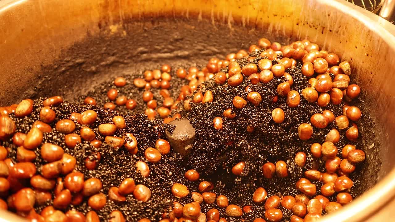 Chestnuts tumble and roast in a large heated wok under warm, golden street food lighting