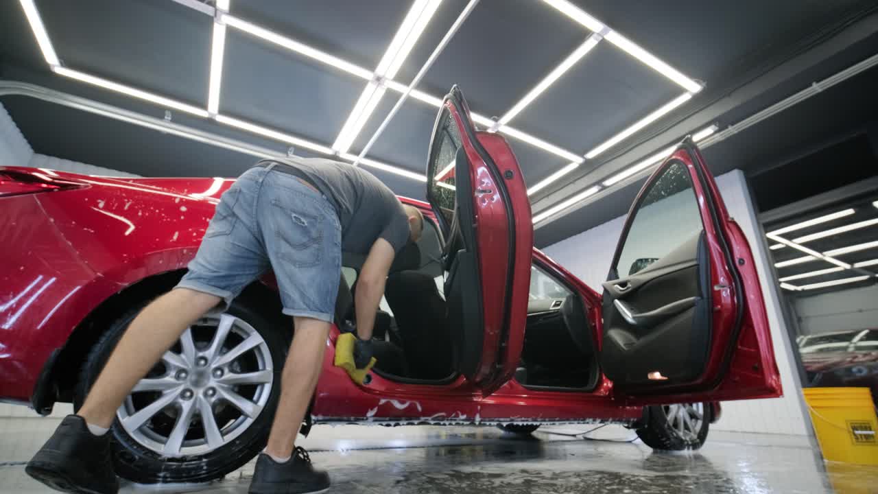 un trabajador lavando un coche rojo en un lavado de coches con un paño amarillo.