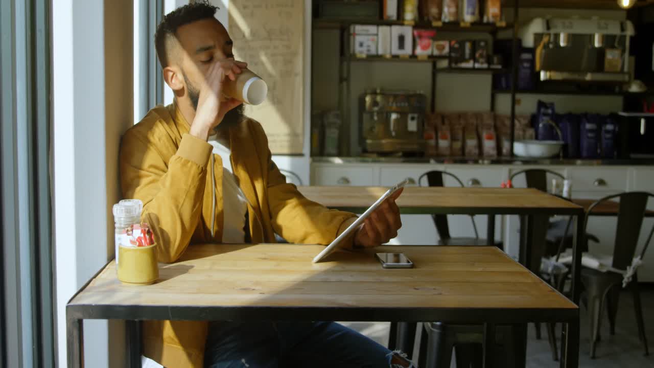 Vista frontale di giovane uomo di etnia mista che beve un caffè e usa un tablet digitale al bar 4K