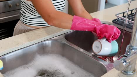 a person wearing pink gloves washes a mug in a well-lit kitchen sink with soapy water
