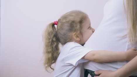 little girl hugs pregnant mother kissing large tummy in room