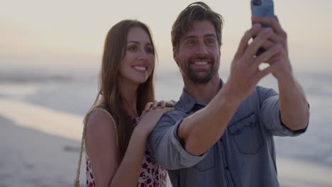 una joven pareja tomando una foto usando un teléfono inteligente disfrutando de una romántica puesta de sol en la playa juntos en cámara lenta