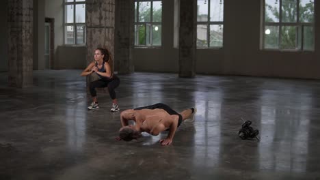 Hermosa-Joven-Pareja-De-Deportes-Está-Trabajando-Juntos-En-El-Gimnasio-Del-Estudio-Con-Ventana-Panorámica.-Una-Chica-Con-Rastas-Hace-Sentadillas,-Un-Hombre-Doblando-Flexiones-Junto-A-Ella