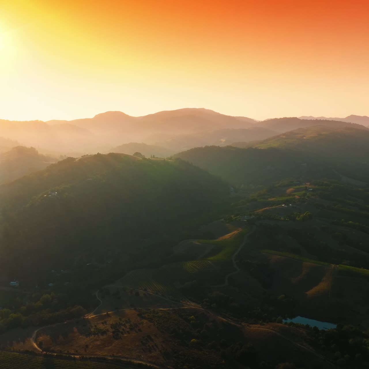 Bright sun lighting the mountainous landscape covered with greenery. Hazy rocky silhouettes at backdrop of pink and orange sky