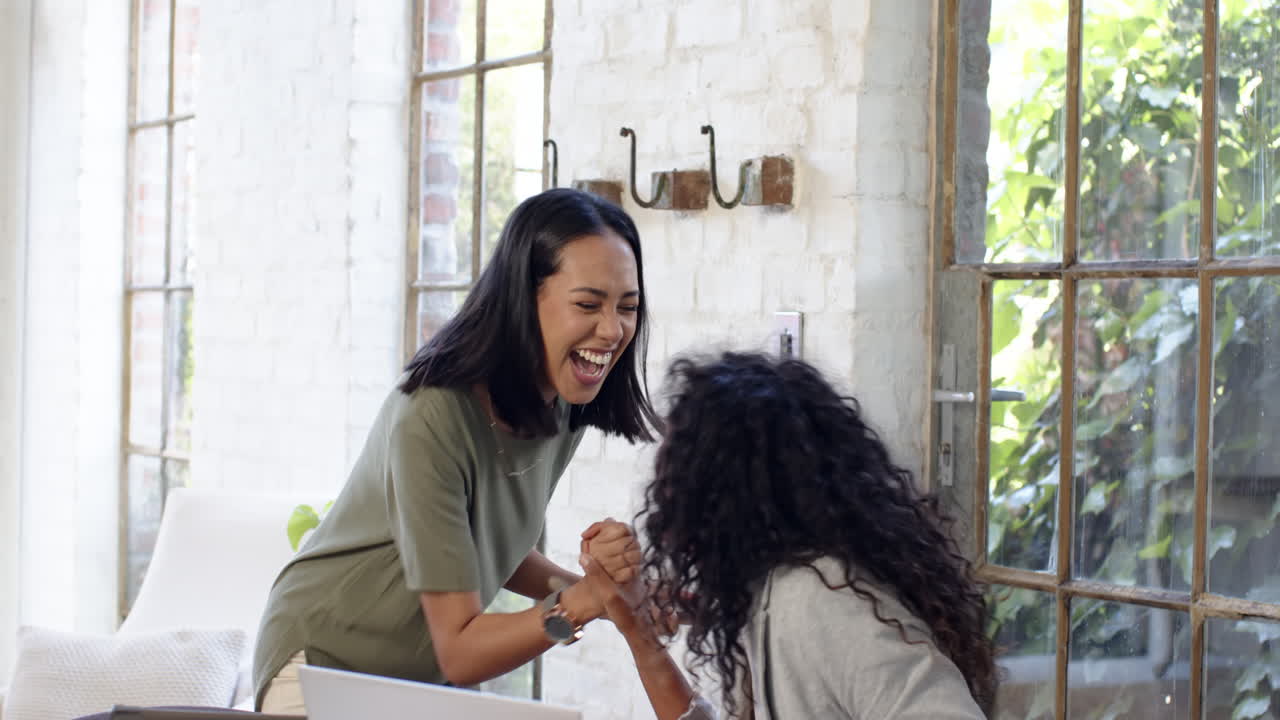 Multiracial female colleagues celebrating at home office, smiling, near smartphone and laptop