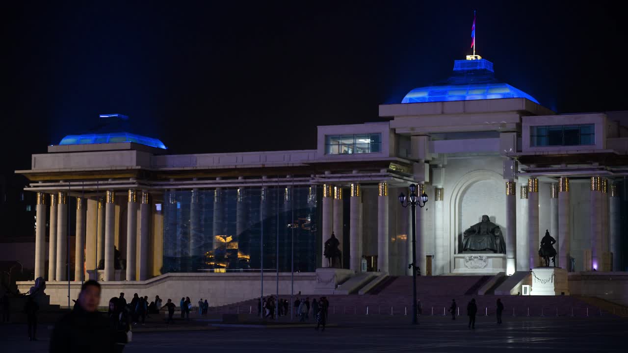 At night, people wander through Sukhbaatar Square in Ulaanbaatar, featuring the bronze statue of Genghis Khan in front of the Government Palace with a flag flying above.