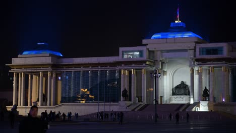 at night, people wander through sukhbaatar square in ulaanbaatar, featuring the bronze statue of genghis khan in front of the government palace with a flag flying above.